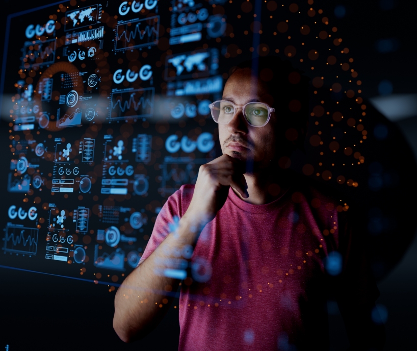 Young man in glasses looks at transparent display showing glowing blue charts.