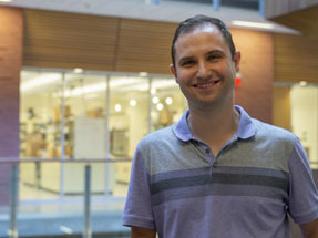 Researcher smiles inside a building with lab rooms in background.